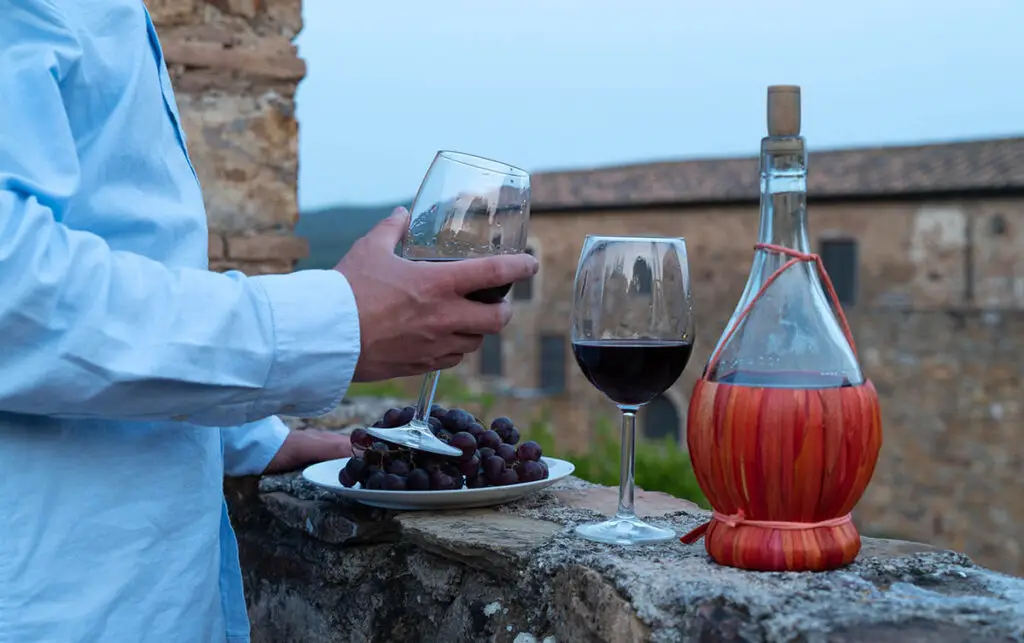 Chianti wine experiences in Tuscany with traditional fiasco bottle, red wine glasses, and grapes on a rustic stone terrace.